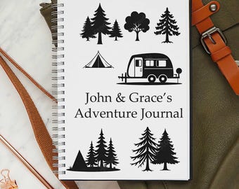 May include: A spiral-bound notebook with a white cover featuring black illustrations of trees, a tent, and a camper. The text "John & Grace's Adventure Journal" is printed in black on the cover. The notebook is lying on a green surface.