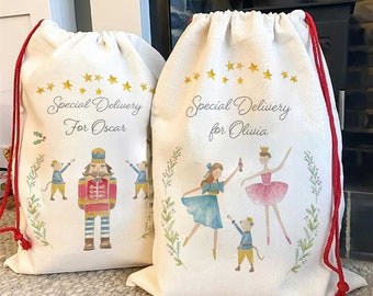 May include: Two off-white drawstring gift bags with red cords. One bag features a Nutcracker and mice, the other a ballerina and dancers. Both bags have the text "Special Delivery" and a name, with star and floral accents.