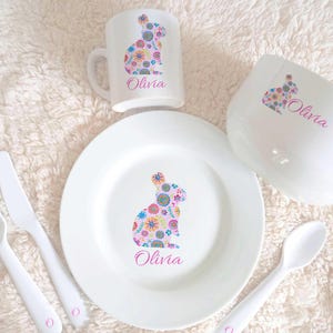 May include: A white children's dinnerware set featuring a floral bunny design. The set includes a plate, bowl, cup, fork, and spoon. The name "Olivia" is printed on each piece.