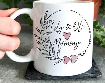 May include: White ceramic mug with a black and white design of a wreath with leaves and a heart. The text "Lily & Oli Mummy" is inside the wreath.