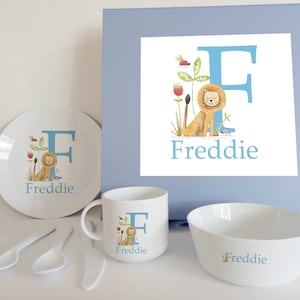 May include: A blue and white personalised children's dinnerware set featuring a lion and the letter F. The set includes a plate, bowl, mug, spoon and fork. The name Freddie is printed on each piece.