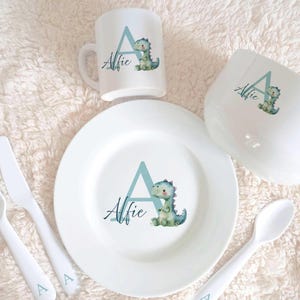 May include: A white children's dinnerware set featuring a blue dinosaur and the letter A. The set includes a plate, bowl, cup, fork, spoon, and a small spoon. The name Alfie is written on each piece.