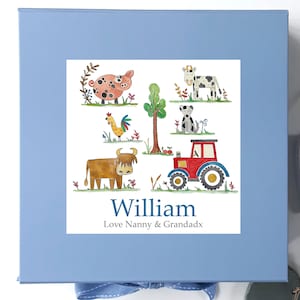 May include: A blue gift box with a white label featuring a farm scene with a pig, cow, dog, rooster, tractor, and a highland cow. The label also includes the name "William" and the text "Love Nanny & Grandadx".