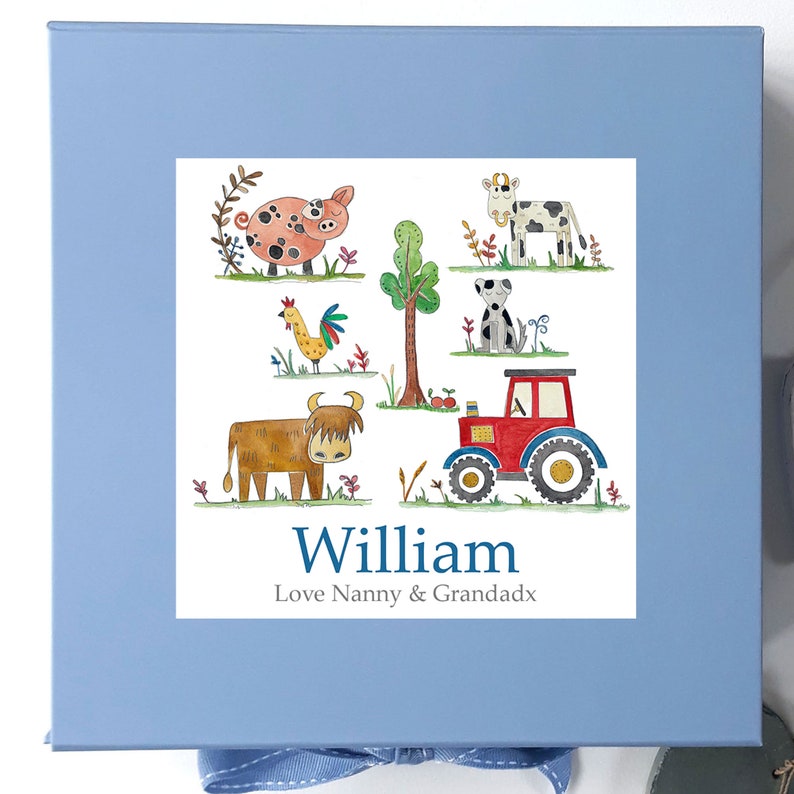 May include: A blue gift box with a white label featuring a farm scene with a pig, cow, dog, rooster, tractor, and a highland cow. The label also includes the name "William" and the text "Love Nanny & Grandadx".