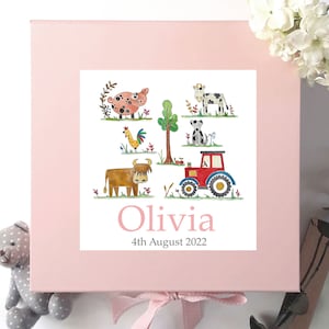 May include: Pink keepsake box with a white label featuring a farm scene with a pig, cow, rooster, dog, tractor, and a brown cow. The label also includes the name Olivia and the date 4th August 2022.