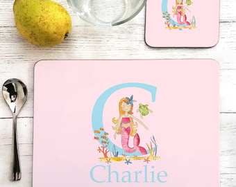 May include: A rectangular pink placemat and a square pink coaster, both featuring a large light blue letter C, a watercolor illustration of a mermaid with a green sea turtle, and the name Charlie printed in light blue on the placemat.