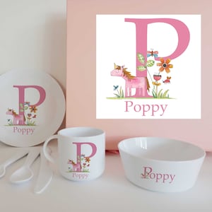 May include: A white children's dinnerware set with a pink letter "P" design, a unicorn illustration, and the name "Poppy". The set includes a plate, mug, and bowl. The unicorn is surrounded by flowers and butterflies.