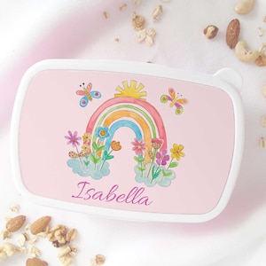 May include: A pink lunchbox with a white trim, decorated with a watercolour illustration of a rainbow, sun, butterflies, and flowers. The name "Isabella" is written in cursive. The lunchbox is rectangular with rounded corners.