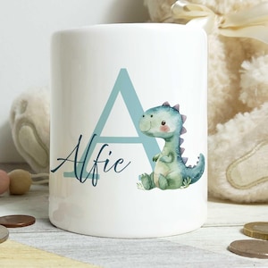 May include: White ceramic money box with a blue letter A and the name Alfie written in cursive. A green dinosaur with a light blue belly is sitting next to the letter A.