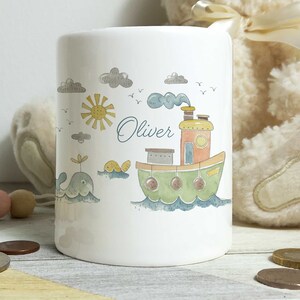 Könnte beinhalten: Weiße Keramik-Sparbüchse mit einem Aquarell-Design eines grünen und orangefarbenen Bootes mit einer gelben Sonne, grauen Wolken und einem blauen Wal. Der Name "Oliver" ist auf der Sparbüchse geschrieben.