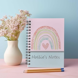 May include: A pink spiral notebook with a watercolour rainbow design and a heart in the centre. The notebook has the text "Hollie's Notes" on the front.