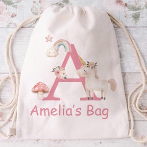 May include: A cream-coloured drawstring bag with a pink letter "A" design, featuring a unicorn, rainbow, cloud, and mushroom. The text "Amelia's Bag" is printed in pink. The bag has a cream-coloured drawstring.