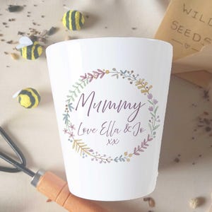 May include: White ceramic plant pot featuring the word "Mummy" within a floral wreath. The pot also includes the text "Love Ella & Jo" and "xx". Decorative bees and a packet of wild seeds are in the background.
