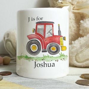 May include: A white ceramic money box featuring a red tractor illustration. The tractor has yellow wheels and a black exhaust pipe. The text 'J is for Joshua' is printed on the box.