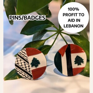 May include: Two round pins or badges with the Lebanese flag design. One pin features the flag with a red, white, and green color scheme. The other pin features the flag with a black and white pattern in the background. The text "100% PROFIT TO AID IN LEBANON" is visible in the image. The text "PINS/BADGES" is also visible in the image.