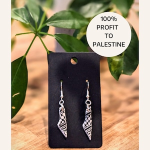 May include: A pair of silver dangle earrings with a black and white patterned design of the outline of Palestine. The earrings are displayed on a black card with the text "100% PROFIT TO PALESTINE".