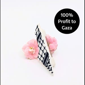 May include: A white and black patterned brooch with a pin backing, resting on a pink flower. The brooch has a geometric design. A black circle with white text reads "100% Profit to Gaza".