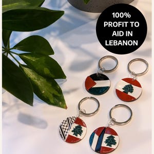 May include: Four keychains with round, colorful, ceramic charms featuring the flag of Lebanon. Each charm has a silver key ring. The text "100% PROFIT TO AID IN LEBANON" is written in black on a white circle.