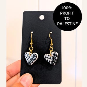 May include: A pair of heart-shaped earrings with a black and white patterned design. The earrings are on a black card with the text "100% PROFIT TO PALESTINE".