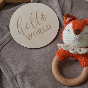 May include: A wooden disc with the words "hello world" engraved on it rests beside a brown baby onesie and a crocheted fox rattle. The fox rattle has an orange body, white face, and wooden ring.