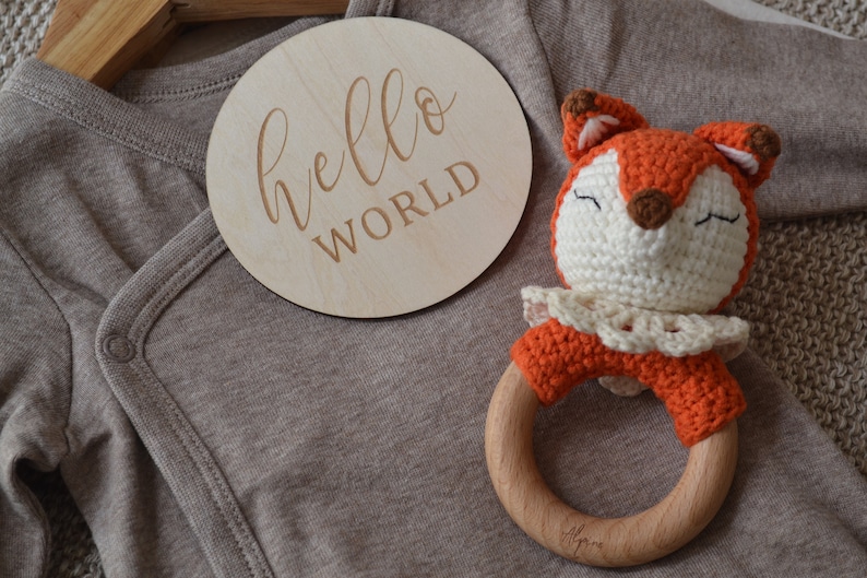 May include: A wooden disc with the words "hello world" engraved on it rests beside a brown baby onesie and a crocheted fox rattle. The fox rattle has an orange body, white face, and wooden ring.