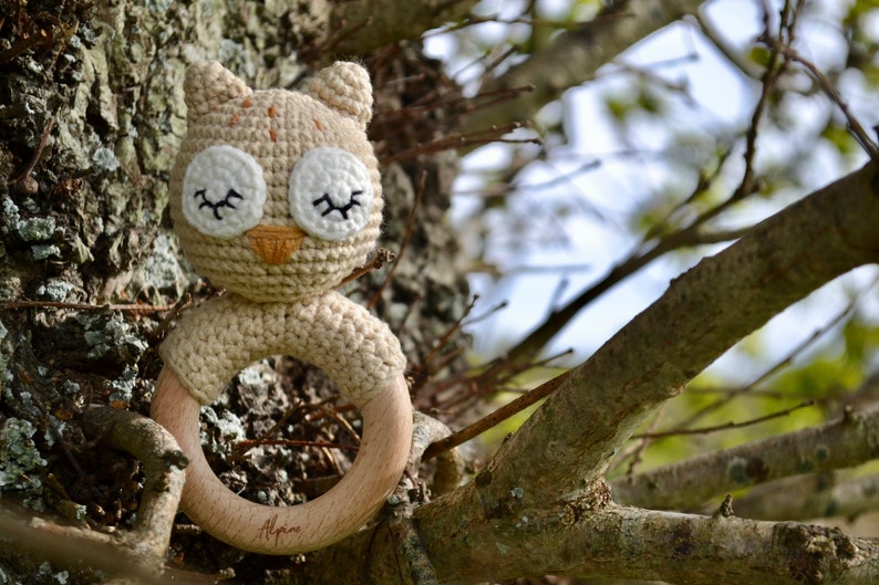May include: A crocheted owl baby rattle with a wooden ring. The owl has white eyes with black eyelashes, a tan beak, and a beige body. The wooden ring is engraved with the word "Alpine".