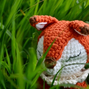 May include: A handmade crochet fox toy with orange, white, and brown details, nestled in tall green grass. The fox has closed eyes, suggesting a peaceful or sleeping pose. The craftwork is visible.