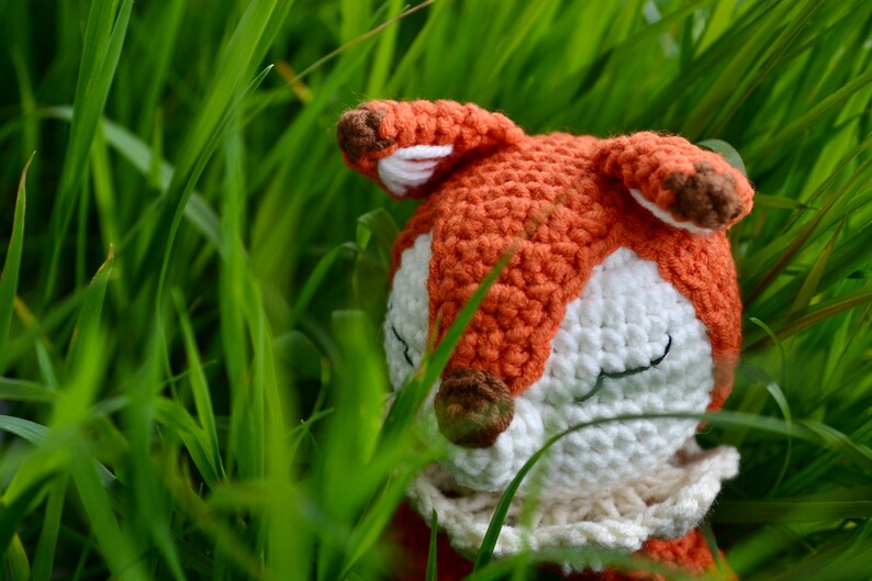 May include: A handmade crochet fox toy with orange, white, and brown details, nestled in tall green grass. The fox has closed eyes, suggesting a peaceful or sleeping pose. The craftwork is visible.