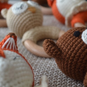 May include: Close-up of several handmade crochet baby rattles. The rattles feature animal designs, including a fox, owl, and bear, with wooden ring handles. The crochet animals are in shades of brown, orange, and white.