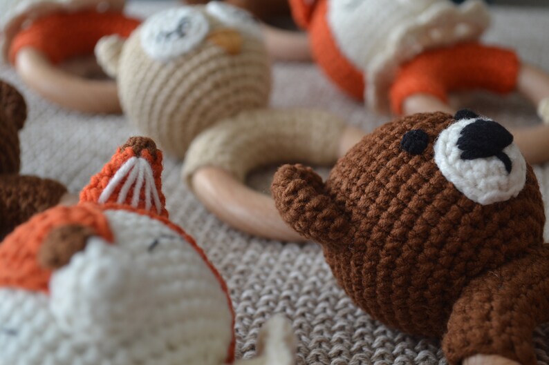 May include: Close-up of several handmade crochet baby rattles. The rattles feature animal designs, including a fox, owl, and bear, with wooden ring handles. The crochet animals are in shades of brown, orange, and white.