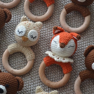 May include: A collection of handmade crochet baby rattles in various animal designs. The rattles feature a wooden ring and are adorned with crochet bears, owls, and foxes in shades of brown, orange, and cream. The toys are designed for babies.