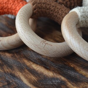 May include: Close-up of three wooden baby teething rings with colorful crochet details. The rings are made of light-colored wood and feature orange, brown, and cream-colored crochet. The word "Alpine" is engraved on the wooden rings.