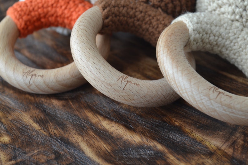 May include: Close-up of three wooden baby teething rings with colorful crochet details. The rings are made of light-colored wood and feature orange, brown, and cream-colored crochet. The word "Alpine" is engraved on the wooden rings.