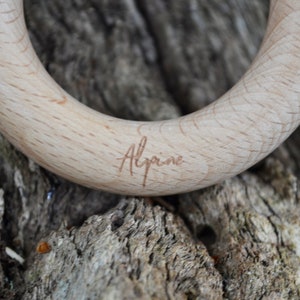 May include: Close-up of a wooden teething ring with the word "Alpine" engraved on the surface. The ring is made of light-colored wood and has a smooth, rounded shape. The background is a textured, natural surface.