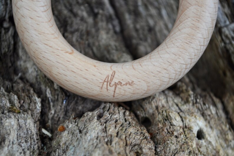 May include: Close-up of a wooden teething ring with the word "Alpine" engraved on the surface. The ring is made of light-colored wood and has a smooth, rounded shape. The background is a textured, natural surface.