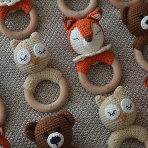 May include: Crocheted baby rattles in the shape of animals, including a fox, owl, and bear. Each rattle features a wooden ring and is made with soft, colorful yarn. The animals have closed eyes, suggesting they are sleeping.