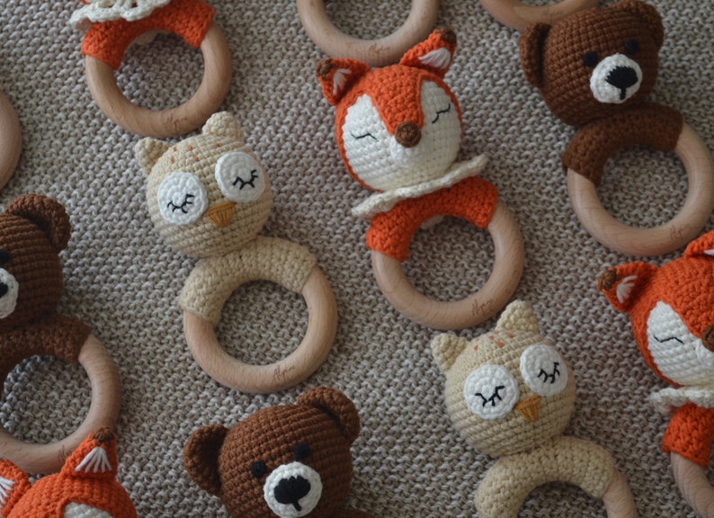 May include: Crocheted baby rattles in the shape of animals, including a fox, owl, and bear. Each rattle features a wooden ring and is made with soft, colorful yarn. The animals have closed eyes, suggesting they are sleeping.
