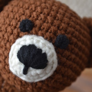 May include: Close-up of a handmade brown crochet bear head. The bear has black eyes and a black nose, with a white muzzle. The crochet stitches are visible, giving it a textured appearance.