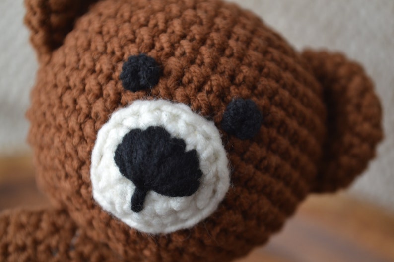 May include: Close-up of a handmade brown crochet bear head. The bear has black eyes and a black nose, with a white muzzle. The crochet stitches are visible, giving it a textured appearance.