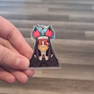 May include: A holographic charm featuring a cartoon character with red hair, a black dress, and a cross on their forehead. The character has a wide, toothy grin and is holding their hands together in a prayer position.