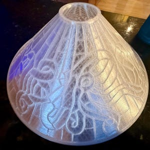 May include: A translucent, conical lamp shade featuring a 3D-printed octopus design with intricate tentacles and a geometric grid pattern. The material is clear and textured, showing a repeating motif of an octopus across the surface of the shade.