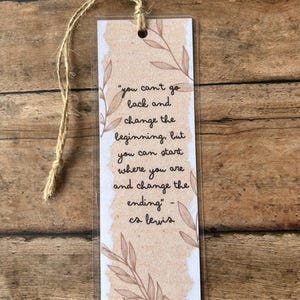 May include: A rectangular bookmark with a quote by C.S. Lewis, featuring a light brown background and floral accents. The quote reads, "you can't go back and change the beginning, but you can start where you are and change the ending." A natural twine is attached.