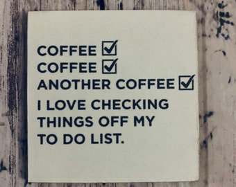 May include: Square, off-white coaster with black text. The text reads "COFFEE ✓ COFFEE ✓ ANOTHER COFFEE ✓ I LOVE CHECKING THINGS OFF MY TO DO LIST."