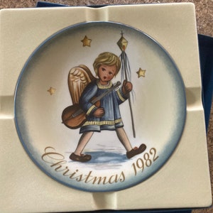 Può includere: Un piatto di Natale in ceramica bianca con un bordo blu. Il piatto presenta un dipinto di un angelo che suona la chitarra e tiene uno stendardo. Il testo "Christmas 1982" è scritto sul piatto.