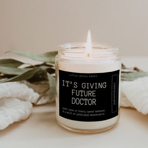 May include: A white candle in a glass jar with a black label that reads "Little Joyful Poppy" and "It's Giving Future Doctor". The label also includes the text "scent notes of freshly opened textbooks & a whiff of caffeinated determination" and "Burn Within Sight".
