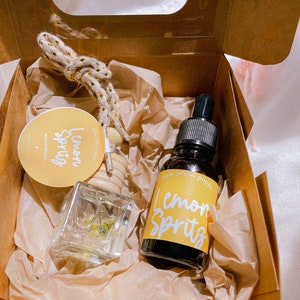 May include: A small brown box with a clear window contains a glass bottle of lemon spritz essential oil and a small glass diffuser with dried flowers inside. The bottle and diffuser have yellow labels with the text "Lemon Spritz" and "little joyful poppy".