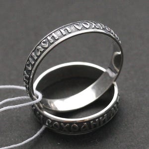 Könnte beinhalten: Zwei silberne Ringe mit graviertem Text. Die Ringe sind gestapelt, wobei einer leicht über dem anderen abgewinkelt ist. Der Text scheint in einer Fremdsprache zu sein. Die Ringe liegen auf einer dunklen Oberfläche.