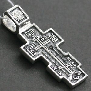 Könnte beinhalten: Ein orthodoxer Kreuzanhänger aus silberfarbenem Metall mit detaillierten Reliefs religiöser Symbole und kyrillischer Schrift. Das Design umfasst ein zentrales Kreuz, einen Speer und ein Schilfrohr sowie ein kleines kreisförmiges Gesichtssymbol.