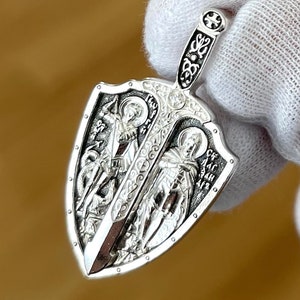 May include: A silver pendant in the shape of a sword and shield. The shield features two religious figures and the sword has an ornate handle. The pendant is held by a gloved hand.