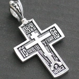 Op de afbeelding: Een orthodoxe kruishanger van zilverkleurig metaal met gedetailleerde in reliëf aangebrachte Cyrillische letters en religieuze iconografie. Het ontwerp bevat een centraal kruismotief, een schedel aan de basis en een decoratief oogje met een klein kruis.